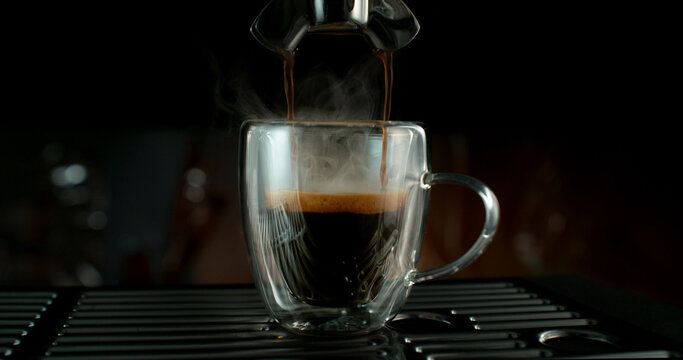 Fresh espresso pouring from coffee machine into glass cup with rich crema and rising steam. Warm cinematic scene capturing preparation of black coffee in cozy atmosphere.