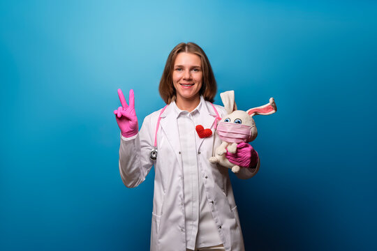 Pediatrician shows the peace sign on a blue background. Pediatrician in a white coat holds a plush toy and showing a peace or two sign with his fingers.