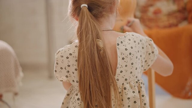 White girl holding pumpkin, cozy kitchen scene with back view of child in floral dress and long ponytail, warm sunlight, rustic table, orange blanket and pillow, intimate autumn baking moment