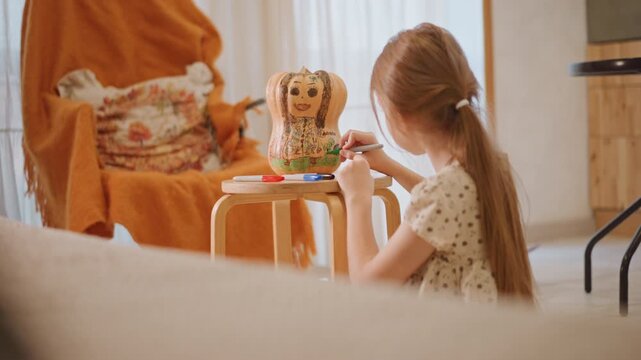 Young white girl decorating pumpkin pie cookie on small table in warm sunlit kitchen, concentrating with paintbrush, cozy armchair and cushion in background, autumn palette, homemade baking