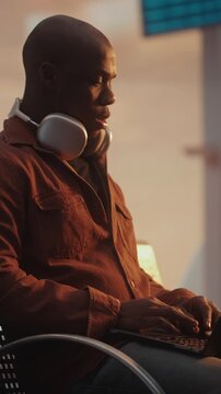 Man Sits on Terminal Bench, Deeply Focused on Laptop Screen While Waiting For Flight. Concept of Freelance Lifestyle, Corporate Travel, Mobile Office Solutions, Global Connectivity. Vertical Shot.