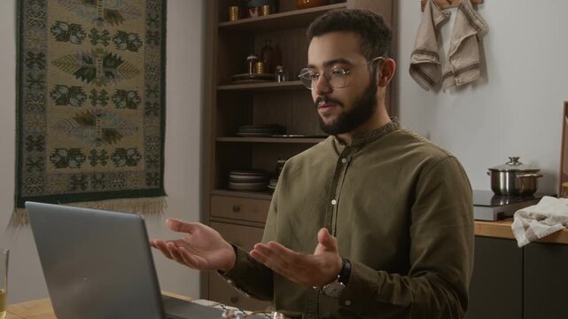 Zoom in shot of modern Muslim young man using laptop in kitchen setting with traditional decor and speaking to colleagues by video chat in online meeting, copy space