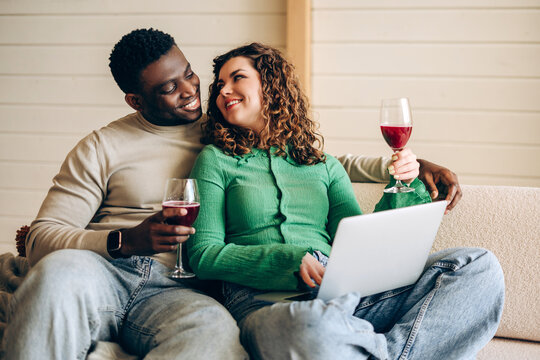 Smiling couple sitting on sofa with laptop and glasses of red wine looking at each other