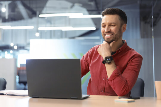 A man is seated at a desk inside an office, looking at a laptop screen. He appears engaged and is smiling while taking part in an online meeting. The office has modern decor.