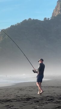 Auckland, New Zealand: A man is surfcasting on a beach with black sand. He is fishing for recreation and to enjoy the outdoors at Karekare Beach.