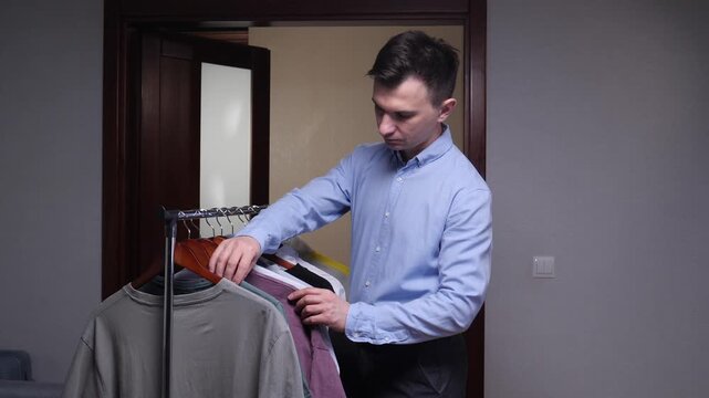 Young man in a modern home selecting clothes from a tidy portable rack, thoughtfully holding a green t shirt as he decides on a casual, comfortable outfit for the day