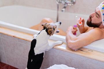 Small dog standing by bathtub while bearded man in shower cap blows bubbles in bright bathroom, creating funny playful mood