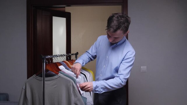 Young man selecting a casual t shirt from a variety of clothes hanging on a portable laundry rack in a modern bedroom, looking for a comfortable outfit