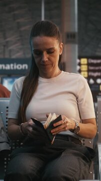 Woman Sits in Departure Hall, Holding Physical Boarding Pass While Checking Details on Smartphone, Having Already Prepared Luggage and Taken Off Coat. Airline Apps, Passenger Security. Vertical Shot.