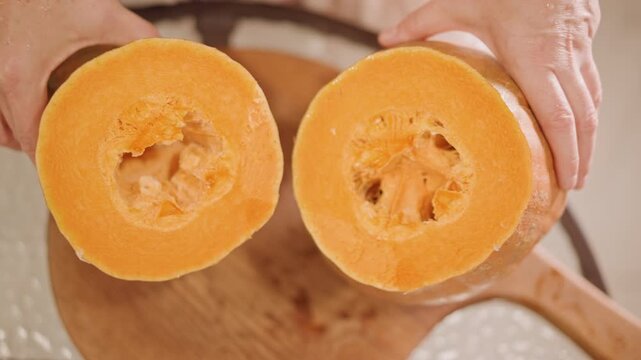 Two halved pumpkins held above board, closeup hands present matching hollowed halves, visible fibrous core and seed pockets, preparation for pie filling or roasting, bright natural light, artisanal