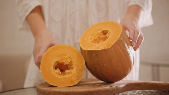 Home cook assembling pumpkin halves. Closeup of hands preparing pumpkin for baking at home. Natural light illuminates hands arranging pumpkin pieces and seeds on rustic kitchen surface