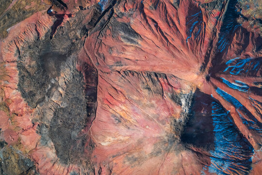 Eroded red slagheap surface with geometric cracks and mineral deposits creating an abstract industrial waste pattern in a detailed drone view.
