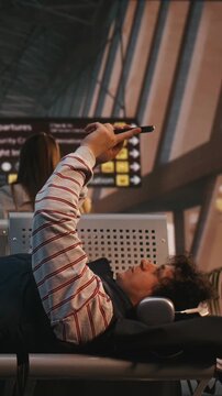 Young Man With Curly Hair, Wearing Large Headphones, Lies Across Row of Airport Chairs in Departure Lounge. Exhausted by Long Wait or Flight Delay, Male Holds Smartphone Above Him, Absorbed in Screen.