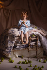 Caucasian girl about 6 in blue dress sitting on stacked mattresses, front view, looking away, beige drapery background, green apples on floor. Concept of fairy tale princess and childhood imagination. © Kaplitskaya Love