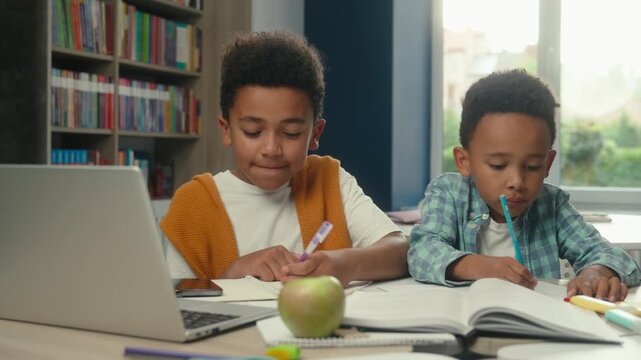 Two African American little boys siblings listening online teacher with laptop writing notes in school library brothers children kids pupils listen tutor virtual course study e-learning write studying