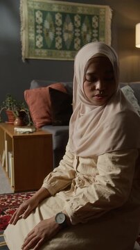 Vertical side view shot of Muslim young woman wearing hijab finishing daily prayer at home and standing up following traditions