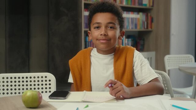 Portrait little African American boy kid child writing exercise math homework son pupil learner studying learning write preparing for exams smiling looking at camera smile at desk in school library