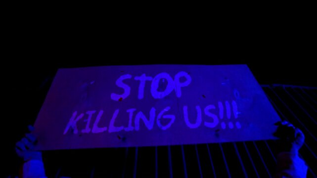 Handheld cardboard sign reading &ldquo;Stop killing us&rdquo; at public protest. Emotional message against violence. Civil unrest and anti violence movement. Social crisis awareness concept.