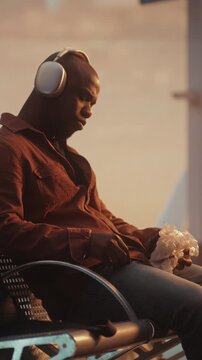 Man Sits on Bench in Airport Departure Lounge, Bathed in Warm Sunset Glow From Panoramic Windows. Male Wearing Headphones and Holds Hamburger, Enjoying Quiet Meal During Transit. Vertical Shot.