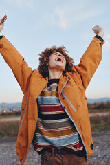 Happy woman with curly hair wearing a rainbow sweater and orange jacket raises arms outdoors. Woman smiles joyfully holding smartphone, lifestyle concept with natural light and open sky background.