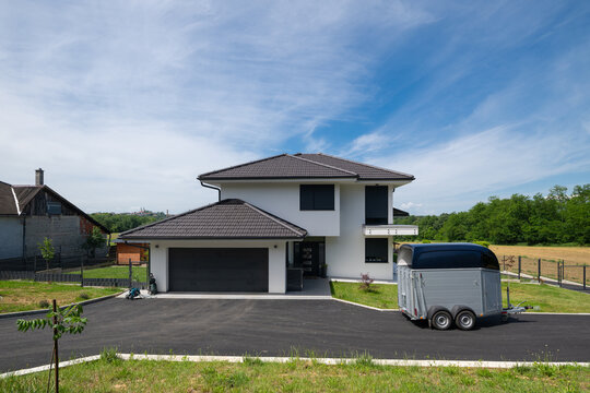 Modern modular minimalist two-story house with garage and trailer on paved driveway in countryside