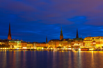 Obraz premium Evening light reflects beautifully across surface of Inner Alster lake in Hamburg. Glowing windows and church steeples line the distant shore of the Binnenalster as the city transitions into night