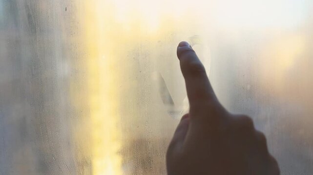 Child's finger is drawing heart shape in circle painted on foggy window covered with drops on sunset