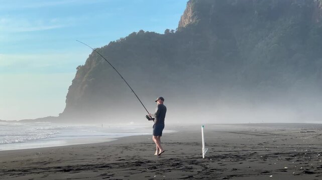 Auckland, New Zealand: A man is surfcasting on a beach with black sand. He is fishing for recreation and to enjoy the outdoors at Karekare Beach.