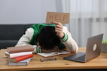 Naklejka na ściany i meble Tired student holding cardboard sign with word Help before exam at wooden desk indoors