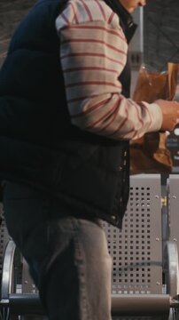 Young Man in Striped Shirt, Navy Vest Sits in Airport Terminal, Enjoying Pastry From Paper Bag. With Backpack on Neighboring Seat, Male Looks Around Slowly While Eating. Travel Moment. Vertical Shot.