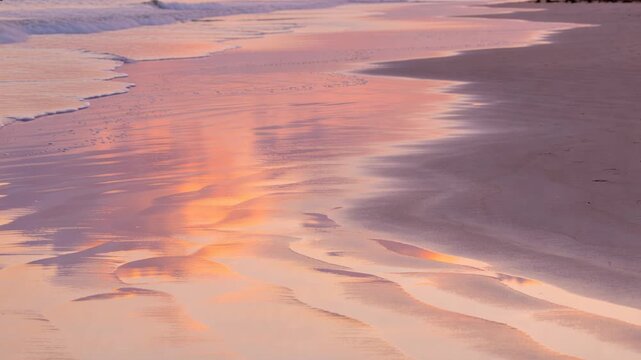 Wet sandy shoreline reflecting warm sky, gentle waves nudging foam and reshaping ripples for travel