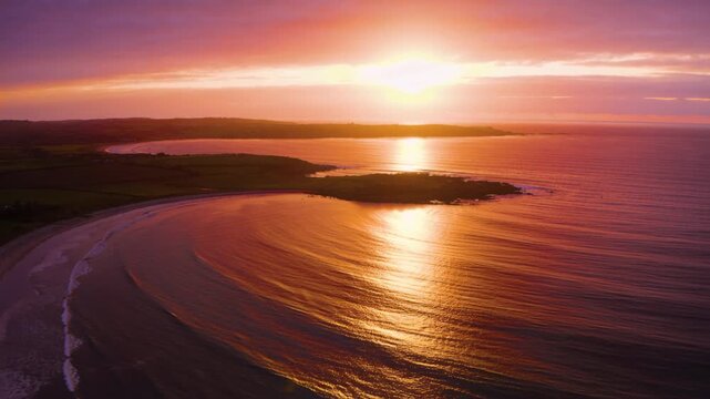 Lowering sun casting golden band across curved sandy bay at sunset, waves advancing and retreating