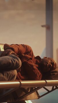 Traveler Lies Down Across Row of Metal Seats in Departure Lounge. Man Uses Backpack As Makeshift Pillow. Fatigue, Discomfort of Long International Transits, Unexpected Flight Delays. Vertical Shot.