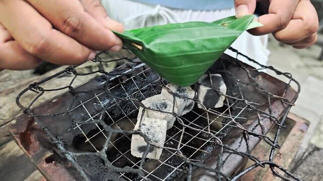 Close-up of a person grilling traditional food called nasi bakar wrapped in banana leaf on a charcoal grill