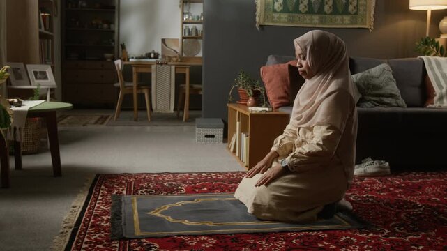 Wide angle shot of young Muslim woman praying at home on prayer mat and performing sujud in cozy home interior with traditional decor, copy space