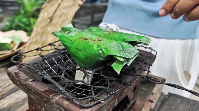 Person grilling traditional banana leaf-wrapped rice, called nasi bakar, on rustic charcoal stove outdoors