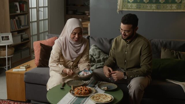 Front view shot of smiling Muslim young woman serving food to husband while enjoying traditional meal together sitting on couch at home