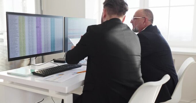 Analysts discuss finance on dual monitors. Two colleagues in suits review spreadsheets, planning budgets and verifying numbers for accurate reporting. Finance teamwork concept clearly presented.