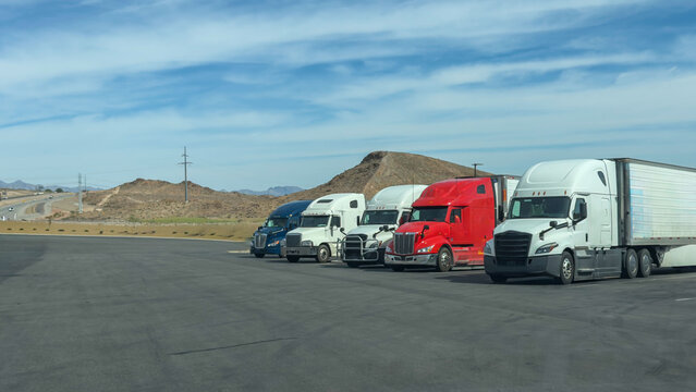 Row of white, gray, red, and black semi-trucks parked parallel at a truck stop. Sunny day, sandy mountains, and blue sky with clouds in the background. American logistics and transport.