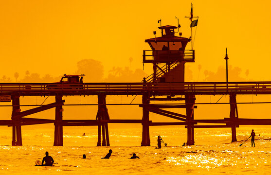 San Clemente Pier at sunset with surfers and paddleboarders silhouetted in golden Pacific Ocean, Southern California