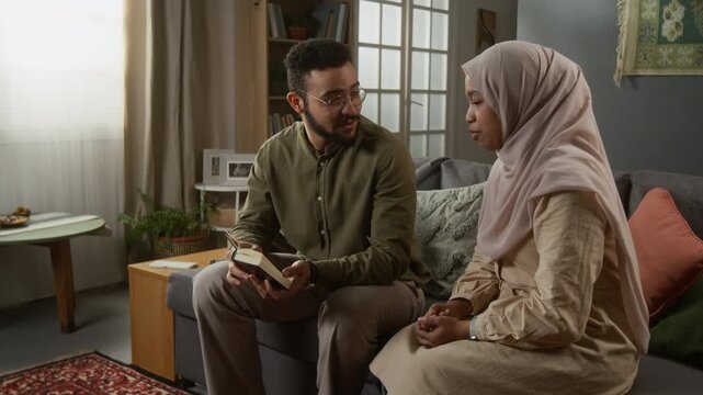 Medium shot of young Muslim couple reading Quran together, man and woman dressed in modest clothing studying religion in cozy home setting