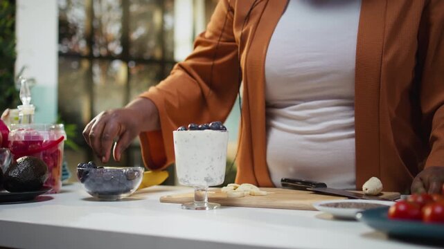 Black woman influencer adding blueberries on a yogurt and chia seeds bowl, creating engaging food vlog content and filming the healthy meal recipe. Chef recommending protein rich breakfast idea.