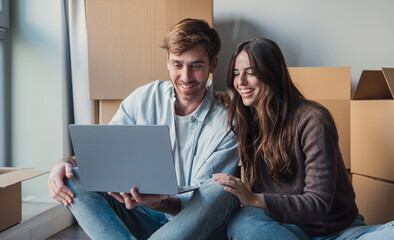 Fototapeta premium A happy young couple sits among cardboard boxes in a bright apartment, smiling and looking at a laptop as they plan, unpack, and organize their new home together on moving day