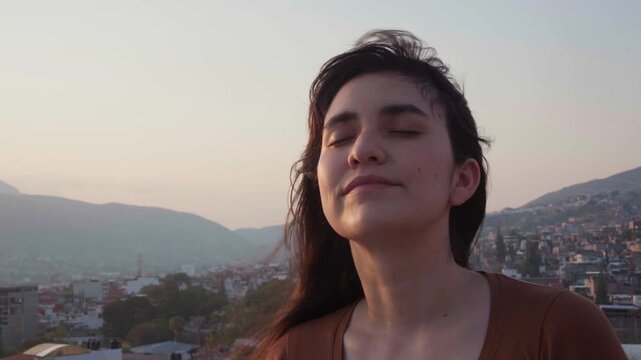 Serene young woman enjoying a tranquil moment while breathing deeply on a rooftop at sunset, feeling the gentle wind in her hair against a beautiful city view