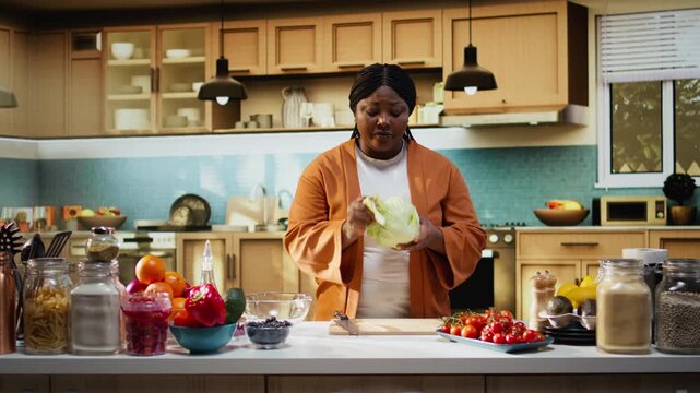 Black food content creator preparing iceberg salad leaves on cooking video, producing online content at home. Solo woman influencer demonstrates meal prep, step by step recipe instructions for vlog.