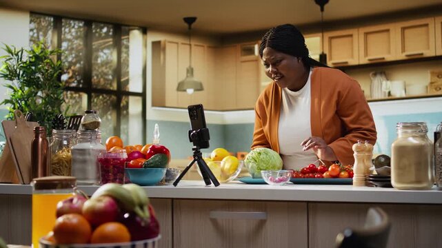 Black food vlogger reading live comments from the audience, recording a livestream on smartphone with tripod as stabilizer in the kitchen. Meal prep cooking vlog for social media.