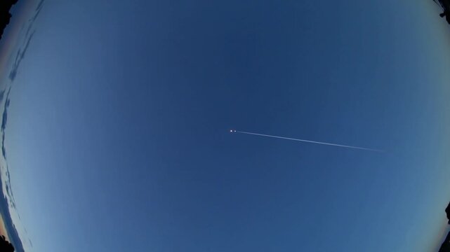 Appearing top-right, twin-lit plane heading right toward horizon in dusk sky with contrail, fisheye
