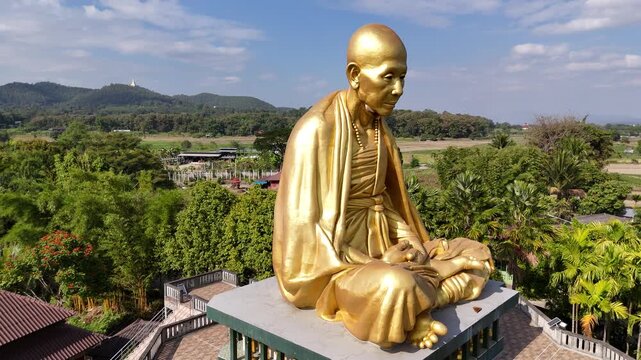 Aerial cinematic view of the giant golden statue of Kruba Srivichai, the patron saint of Lanna, surrounded by mountains in Chiang Rai, Thailand.