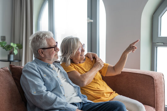Senior couple relax on a couch, smiling as they point toward a bright window and share a tender moment of companionship, affection and contentment in their cozy living room.