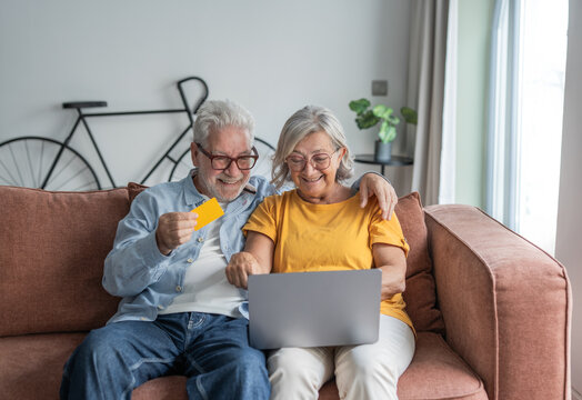 Happy senior couple sits on a comfortable sofa using a laptop and credit card to shop online, sharing a warm moment of connection, convenience and confidence in digital payments at home.
